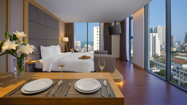 A hotel room with a large bed, a dining table set with plates and cutlery, a vase of flowers, and a city view through large windows. - Estrella Boutique Hotel (Da Nang)