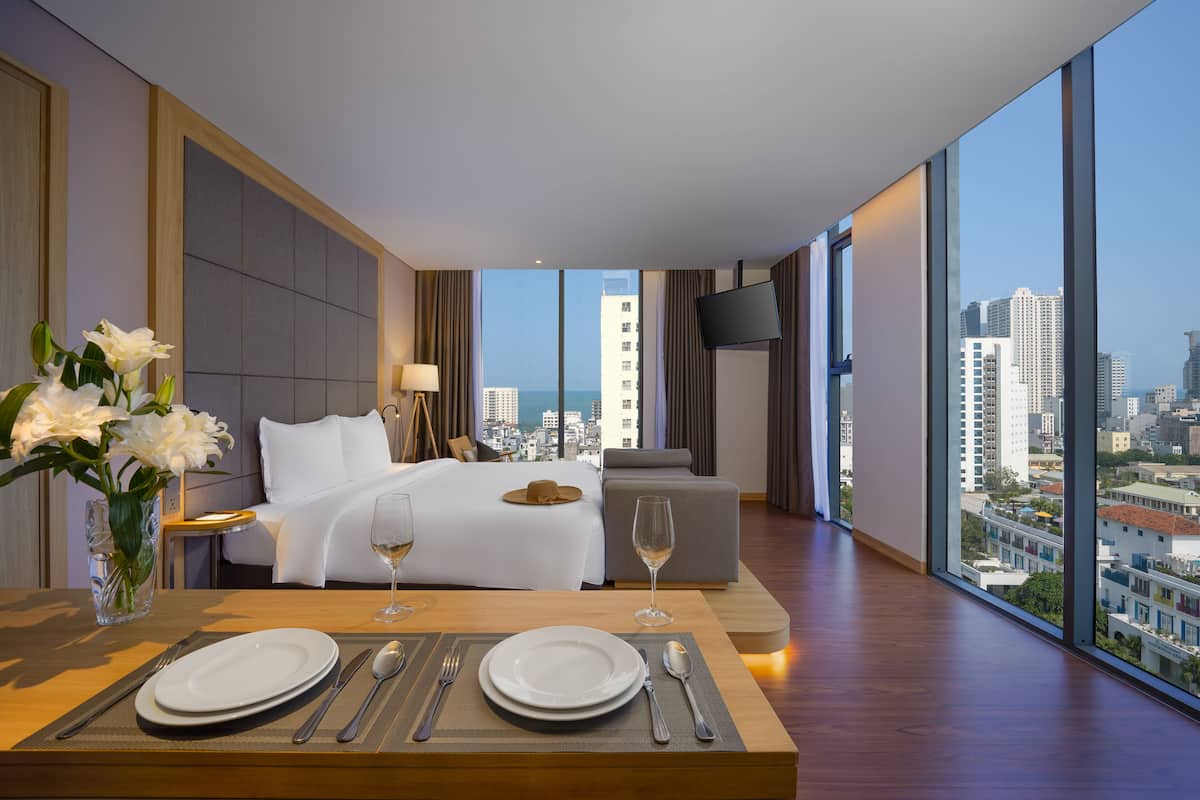 A hotel room with a large bed, a dining table set with plates and cutlery, a vase of flowers, and a city view through large windows.