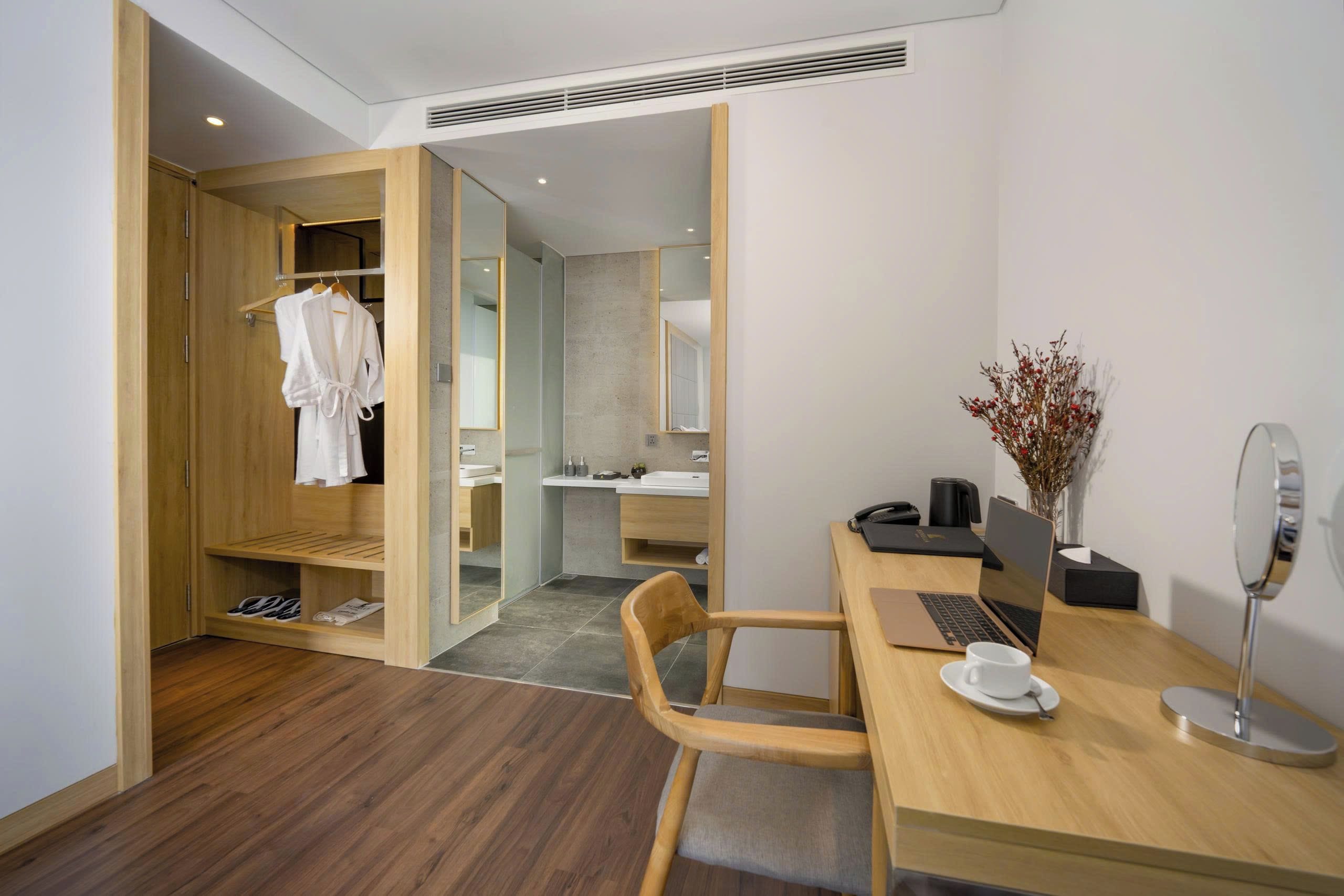A hotel room with a wooden desk, a laptop, a chair, a small table with a cup and saucer, a mirror, and a bathroom visible through an open door.