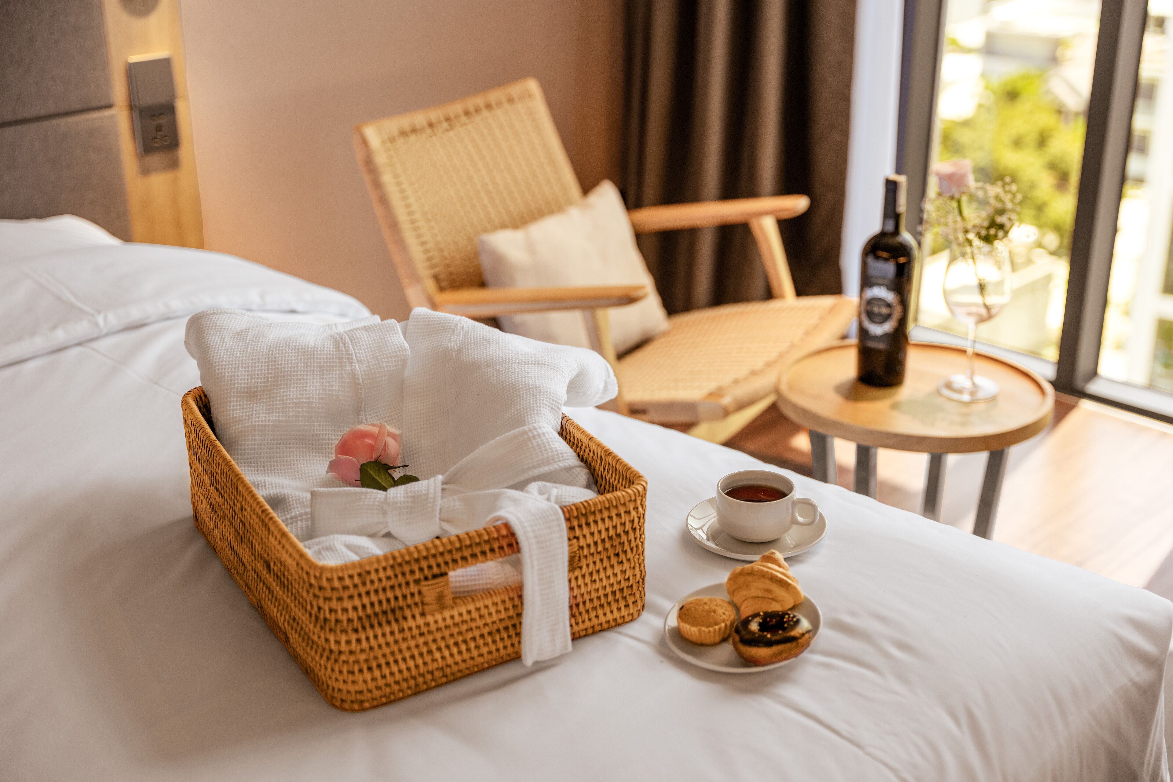 A neatly made bed with a wicker basket containing a rose and a white cloth, a plate of cookies, a bottle of wine, and a glass of wine on a small table beside the bed.
