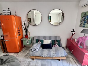 A bedroom with a bed, a dresser, a mirror, a wall-mounted air conditioner, and a vintage orange refrigerator.