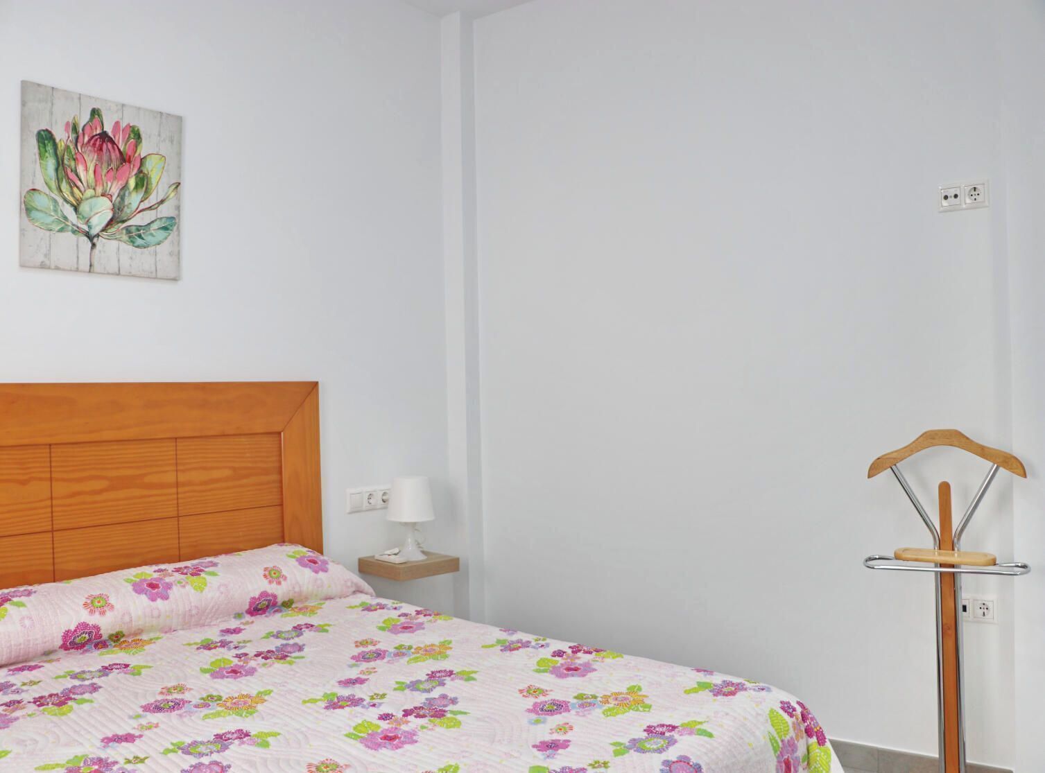 A bedroom with a wooden headboard, a floral bedspread, a bedside table with a lamp, and a wooden hanger on a metal stand.