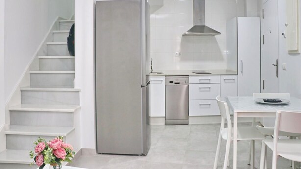 A modern kitchen with white cabinetry, a stainless steel refrigerator, a dishwasher, and a dining area with a table and chairs. - Junquera Apartment in Conil (Conil)