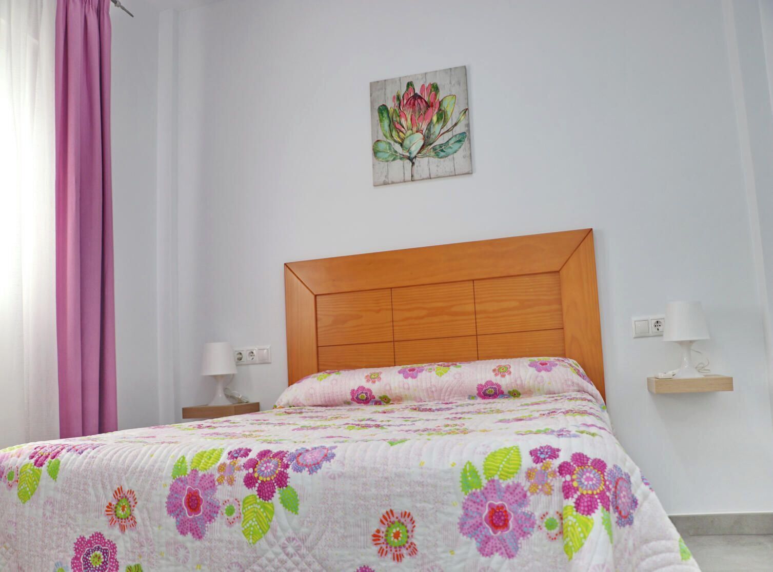 Un dormitorio con cabecera de madera, colcha con estampado floral, mesita de noche con lámpara y un enchufe en la pared.