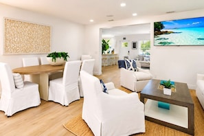 A modern living room with a dining area, white furniture, a large flat-screen TV, and a wall-mounted artwork.