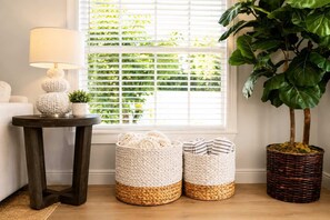 A room with a dark wooden side table, a lamp, two woven baskets, a large potted plant, and a window with blinds.