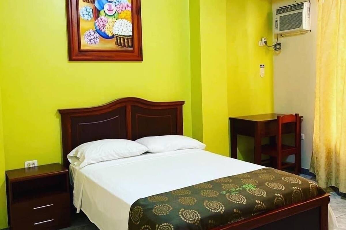 A hotel room with a bed, a nightstand, a small table, and an air conditioning unit.