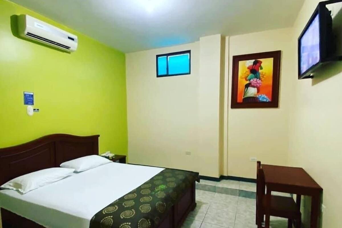 A hotel room with a bed, a small table, a chair, an air conditioner, a window, and a television.