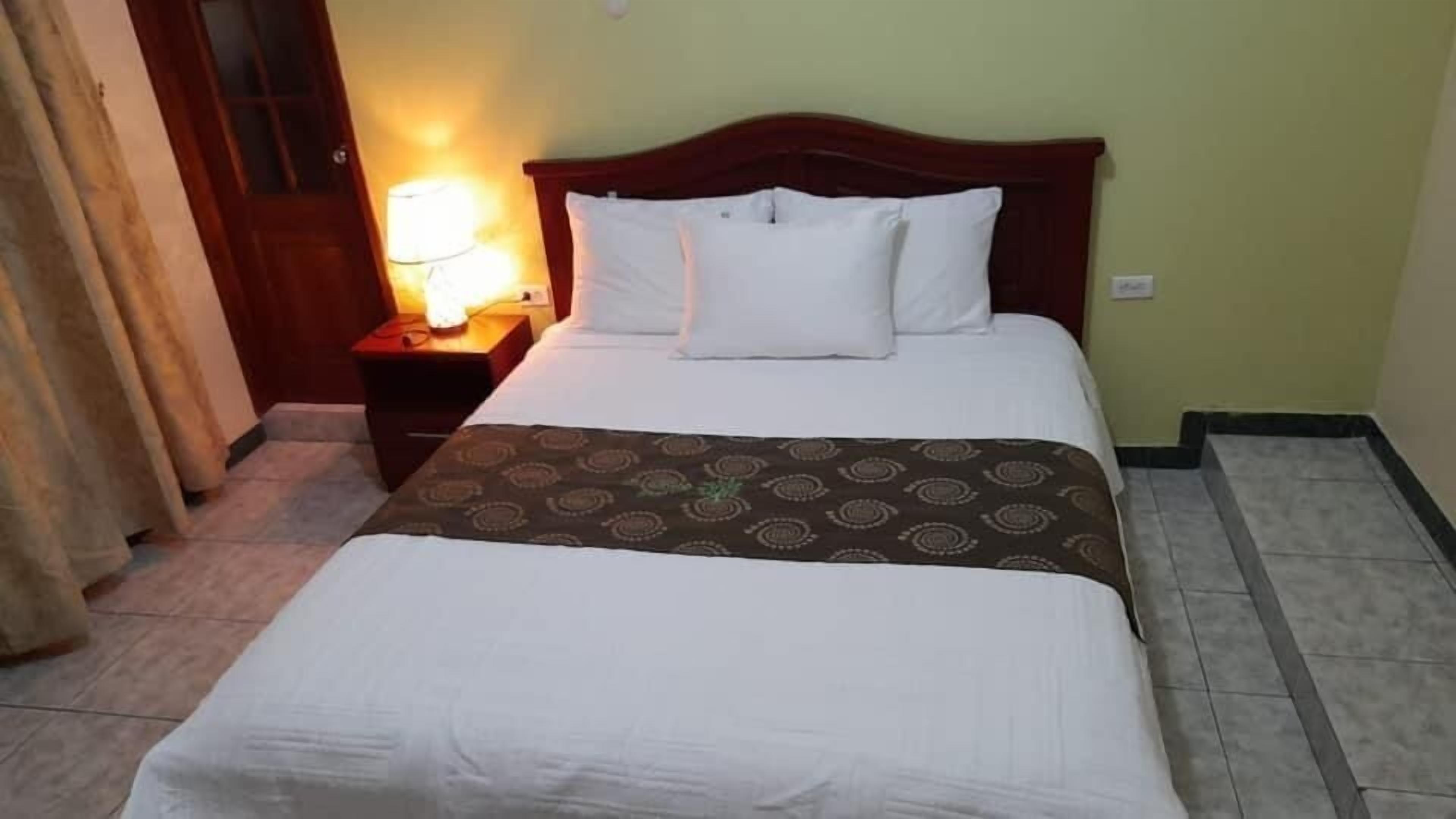 Una habitación de hotel con cama, mesita de noche, lámpara y cortina.