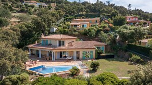 Una amplia villa con piscina, rodeada de exuberante vegetación y otras casas en una ladera.