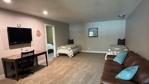 Une chambre dotée d’un téléviseur à écran plat, d’un bureau en bois, de deux lits avec une literie à motifs, d’un canapé en cuir marron et d’une petite fenêtre.