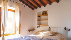 A bedroom with a bed, a window with curtains, a wooden ceiling, and a wall-mounted shelf.