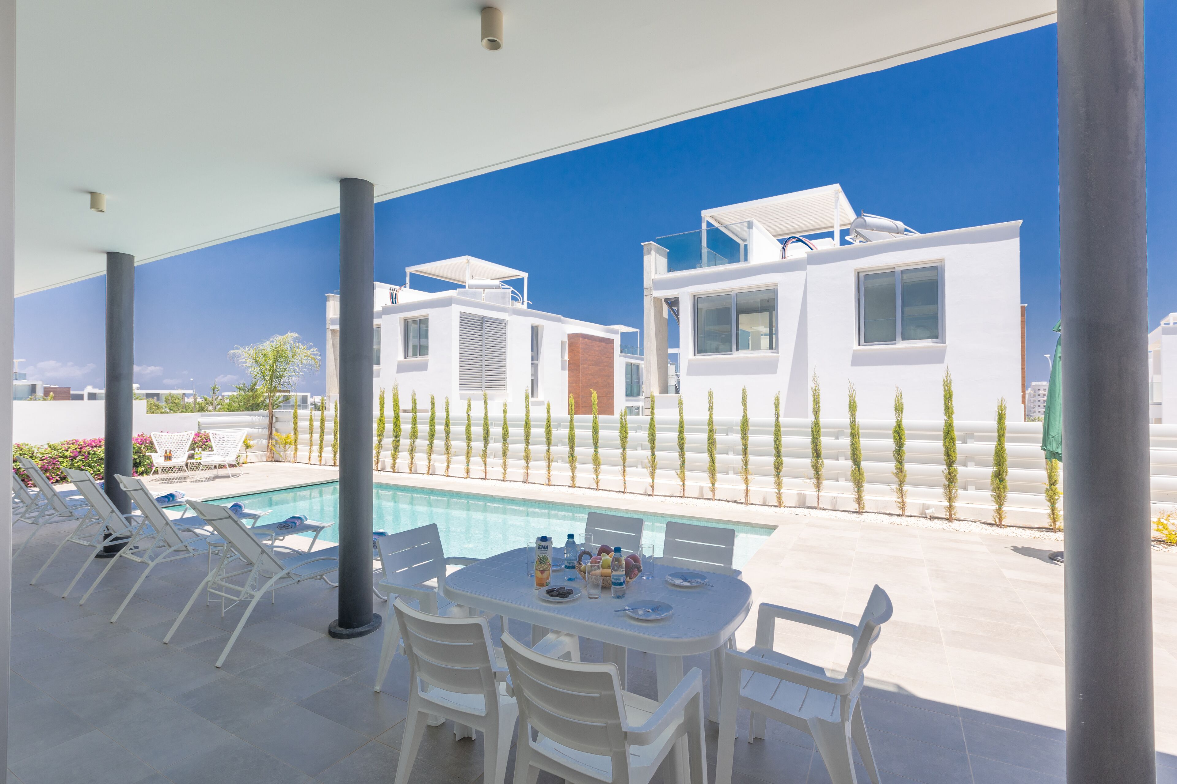 A modern outdoor pool area with white lounge chairs and a table set for dining, surrounded by a clear blue sky and a view of contemporary buildings.