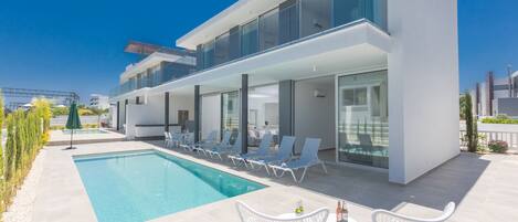A modern building with a swimming pool, lounge chairs, and a table with drinks.