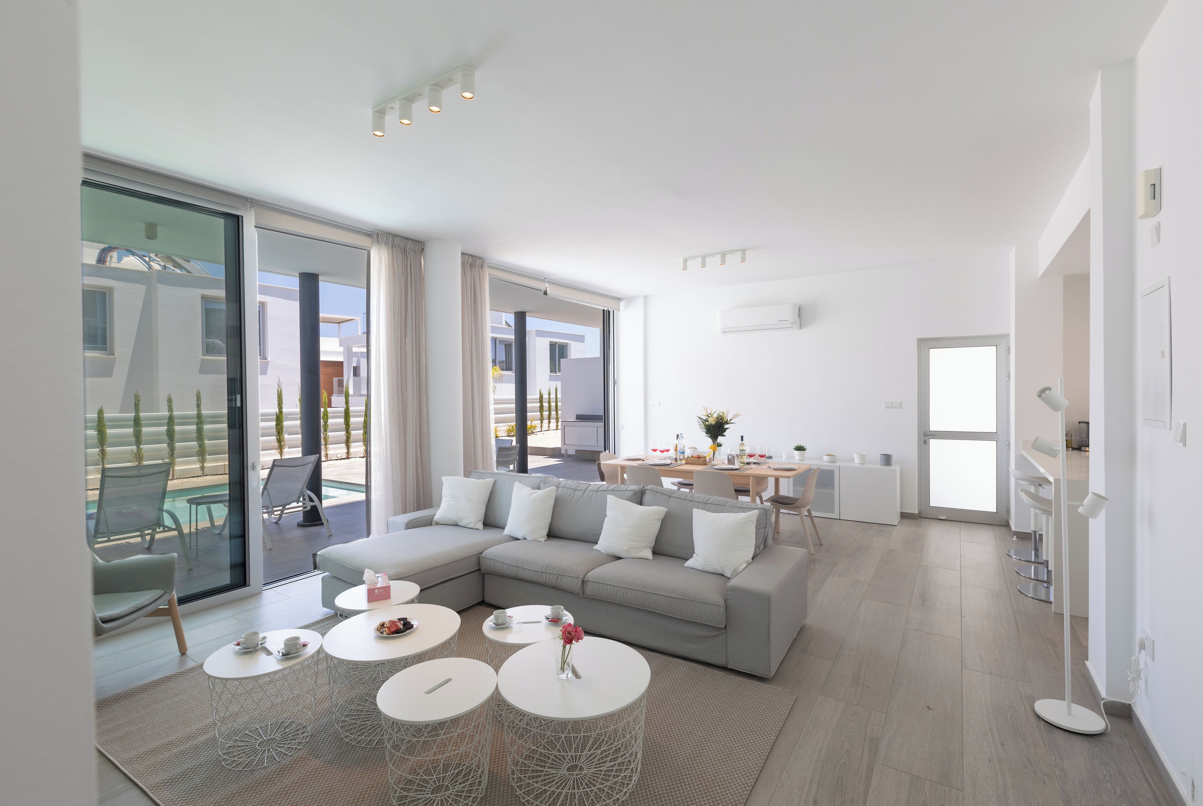 Una sala de estar moderna con un sofá seccional gris, mesas de centro redondas blancas y una puerta corredera de vidrio que da a un espacio exterior.