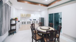 A dining area with a round table and chairs, a kitchen in the background, and a sliding glass door leading to an outdoor area. - Paradise Found 4BR Villa,Fits 22 PAX, 6-Min to Lee Garden (Hat Yai)