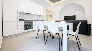 A modern kitchen with white cabinetry, a dining table with chairs, and a black sofa in a living area with a TV.