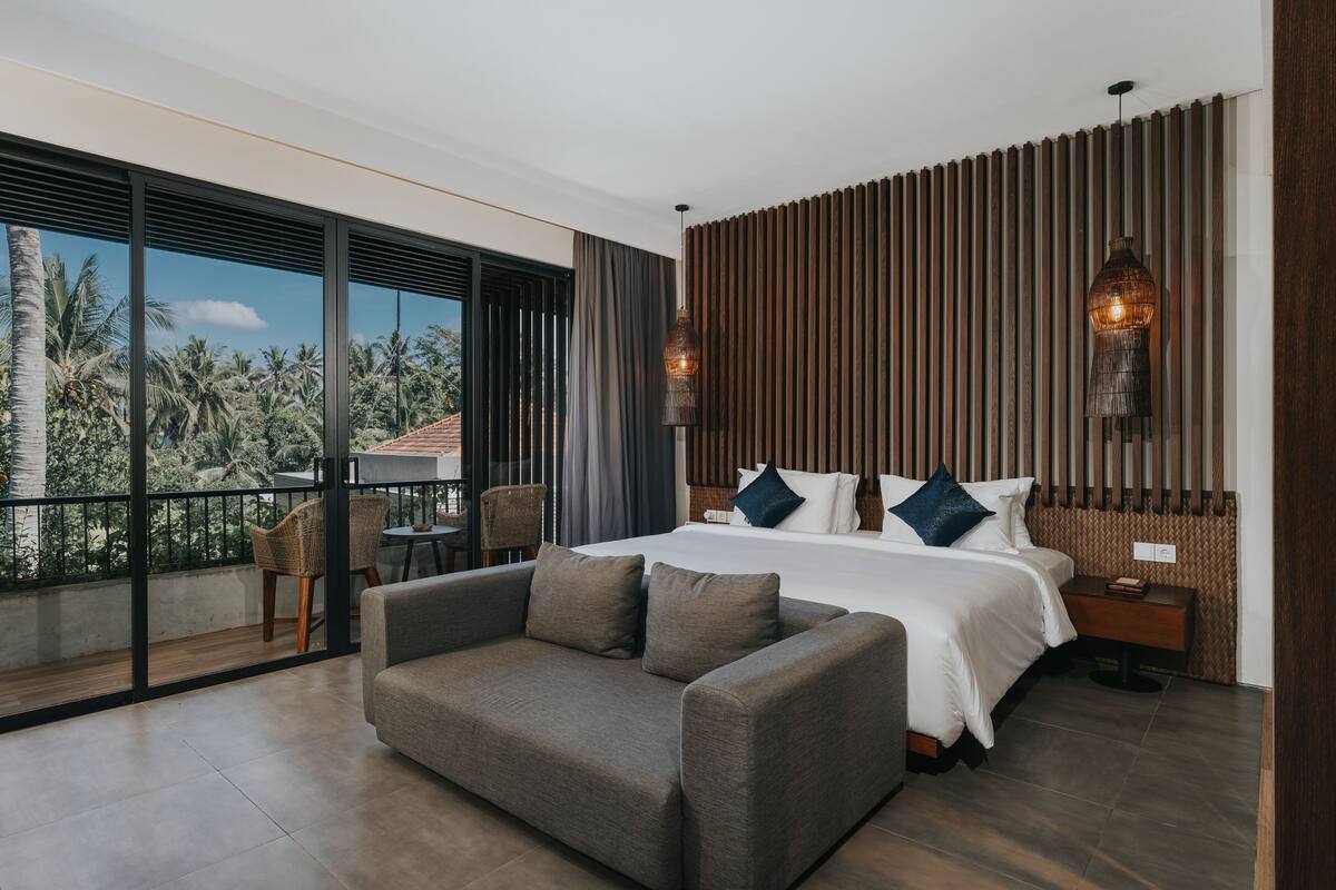 A modern hotel room with a large bed, a sofa, a balcony with a view of palm trees, and a wooden wall panel.