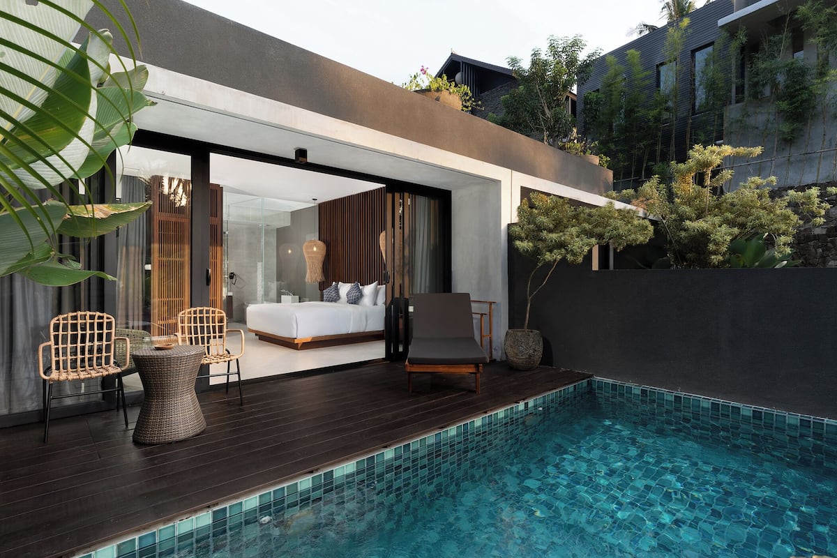 A modern outdoor pool area with a wooden deck, lounge chairs, and a glass-enclosed bedroom with a bed and seating area.