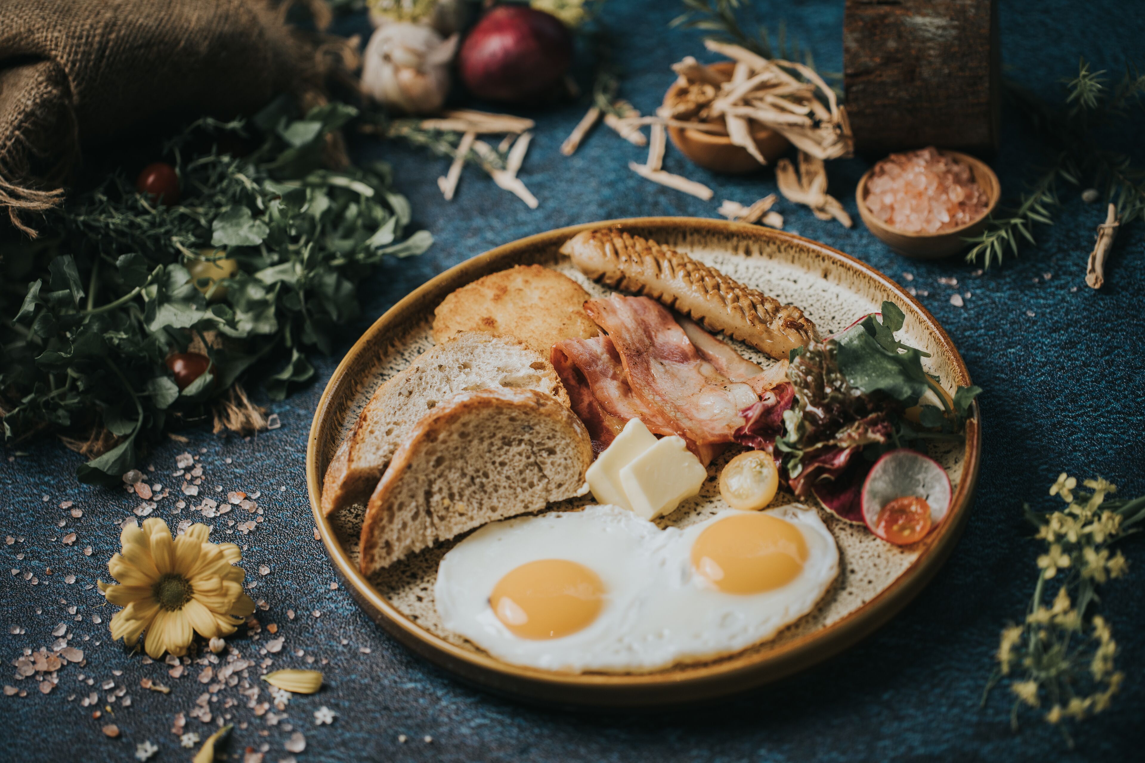 A breakfast plate with eggs, bacon, toast, and greens on a textured blue surface.