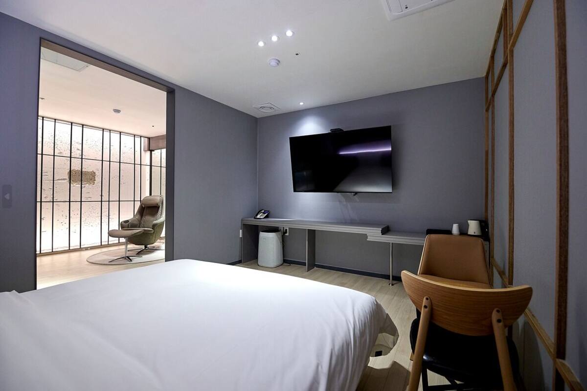 A modern hotel room with a large bed, a TV mounted on the wall, a desk with a chair, and a window with a view of the beach.
