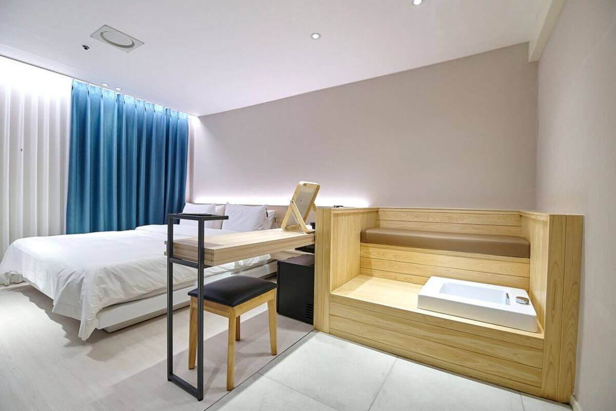 A modern hotel room with a bed, a desk with a chair, a wooden bench with a sink, and a large window with curtains.