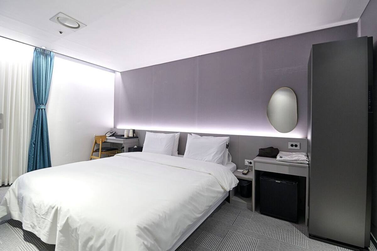 A modern hotel room with a large bed, a desk, a small refrigerator, and a wall-mounted mirror.