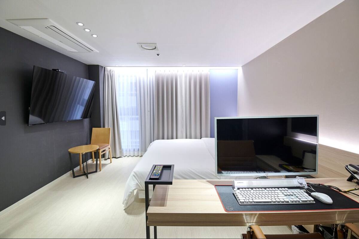 A modern hotel room with a bed, a desk with a computer, a TV, and a chair.