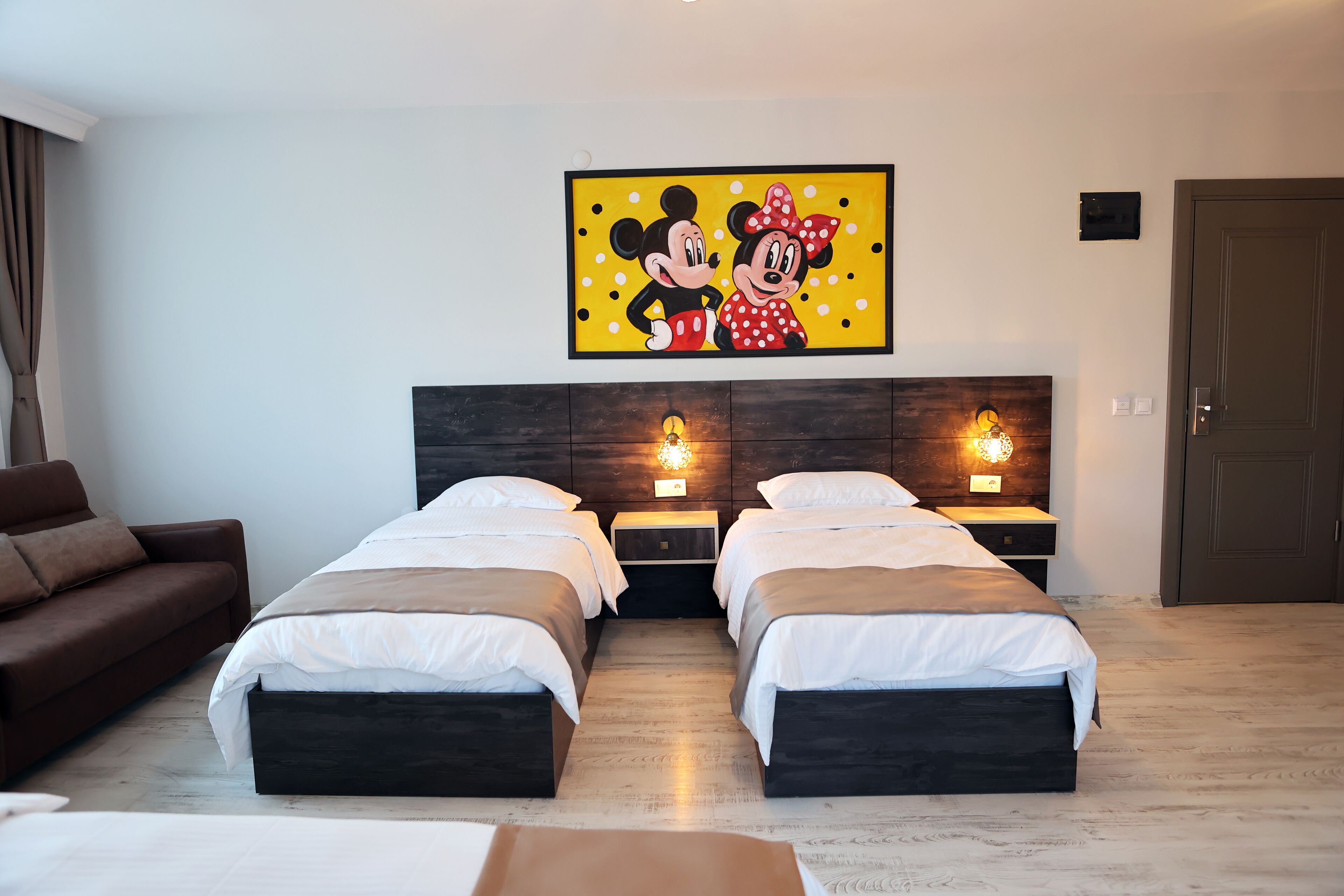 A hotel room with two beds, a brown sofa, a wall-mounted picture of Mickey and Minnie Mouse, and bedside lamps.