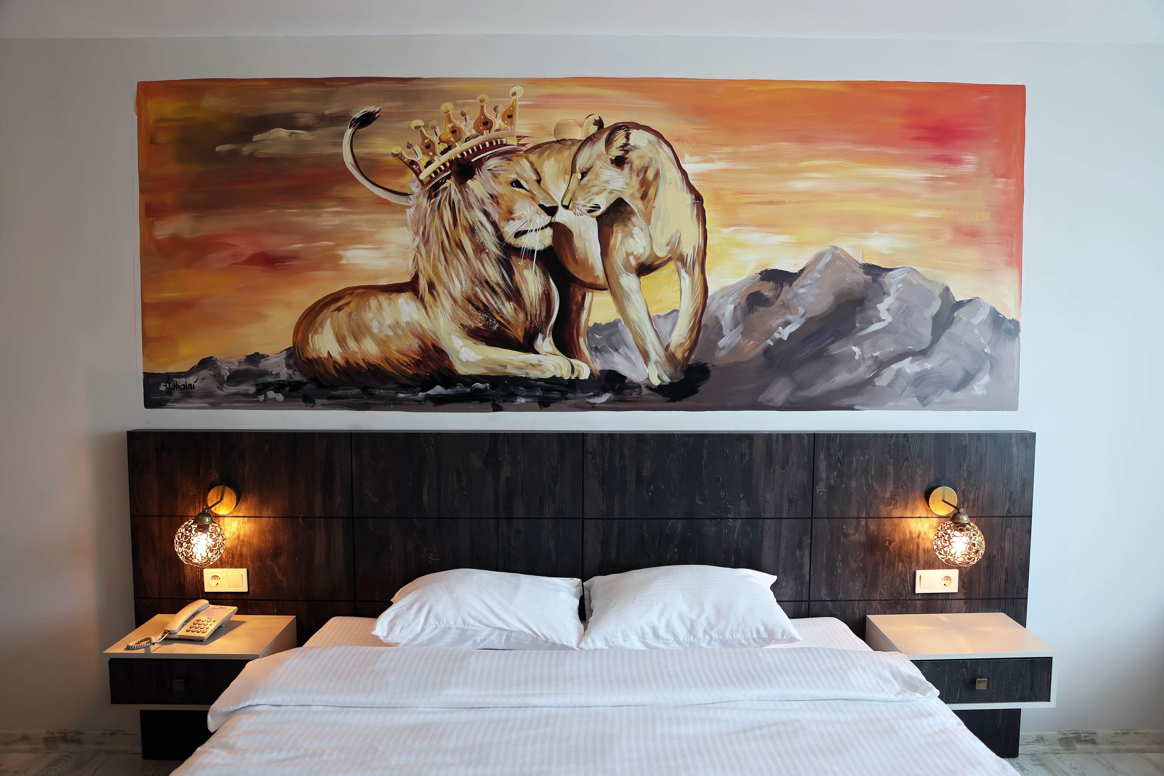 Habitación de hotel con un cuadro grande de leones, una cama con sábanas blancas, dos mesitas de noche con lámparas y un cabecero de madera.