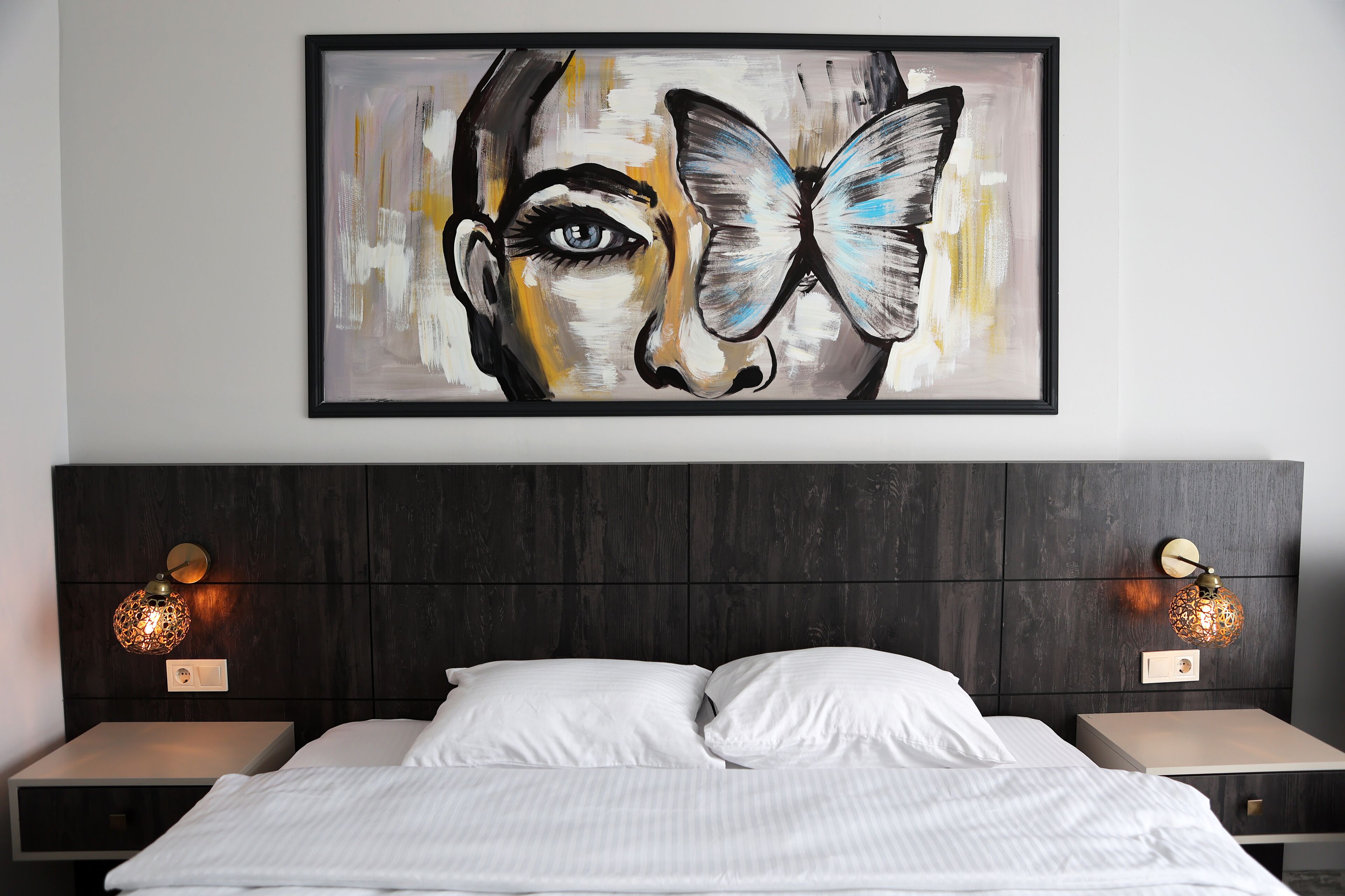 A bedroom with a large framed painting, a dark wood headboard, two bedside tables with lamps, and a bed with white linens.
