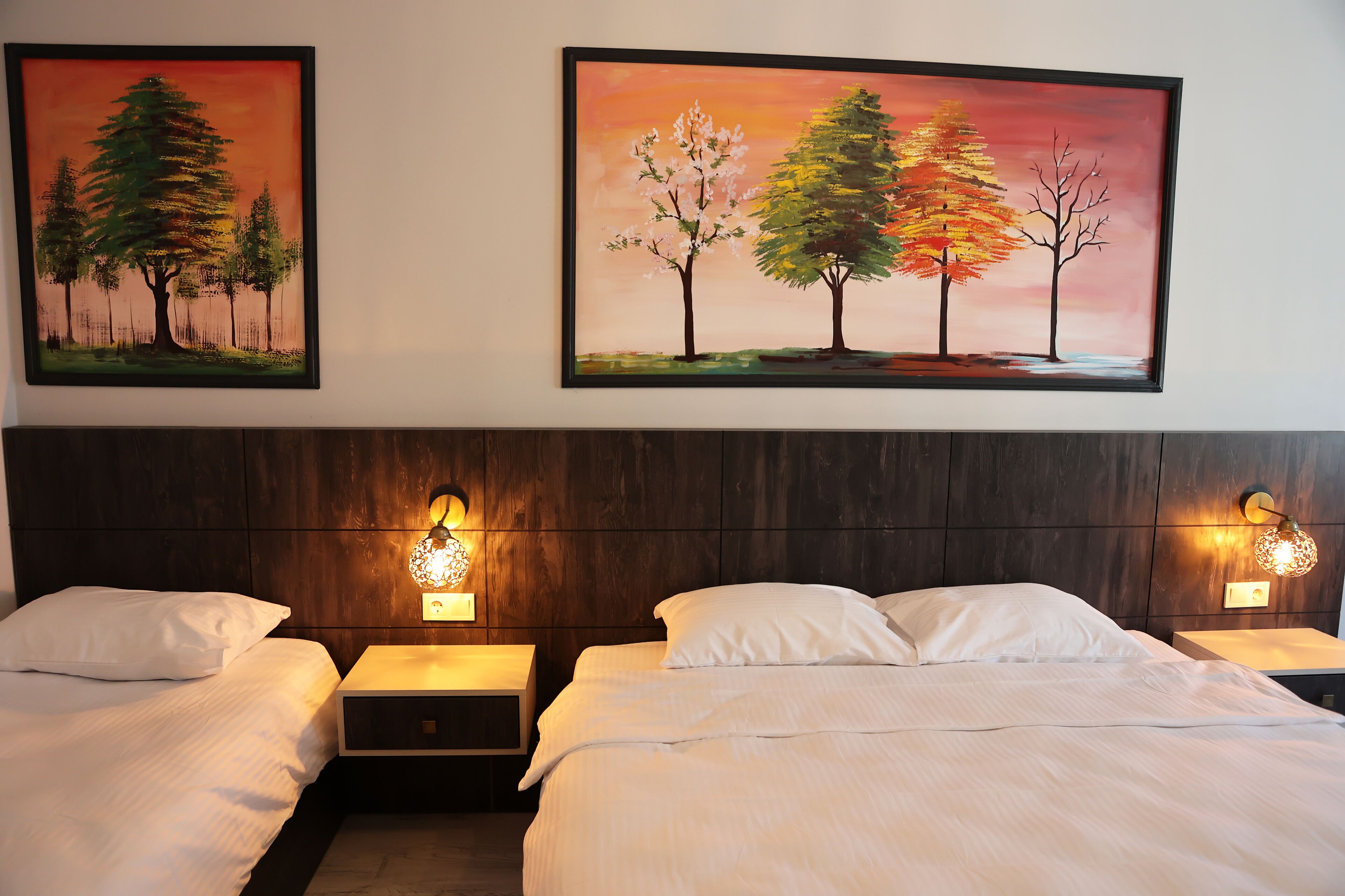 Une chambre d’hôtel avec deux lits, une tête de lit en bois, des tables de chevet avec des appliques murales et deux tableaux encadrés représentant des arbres.