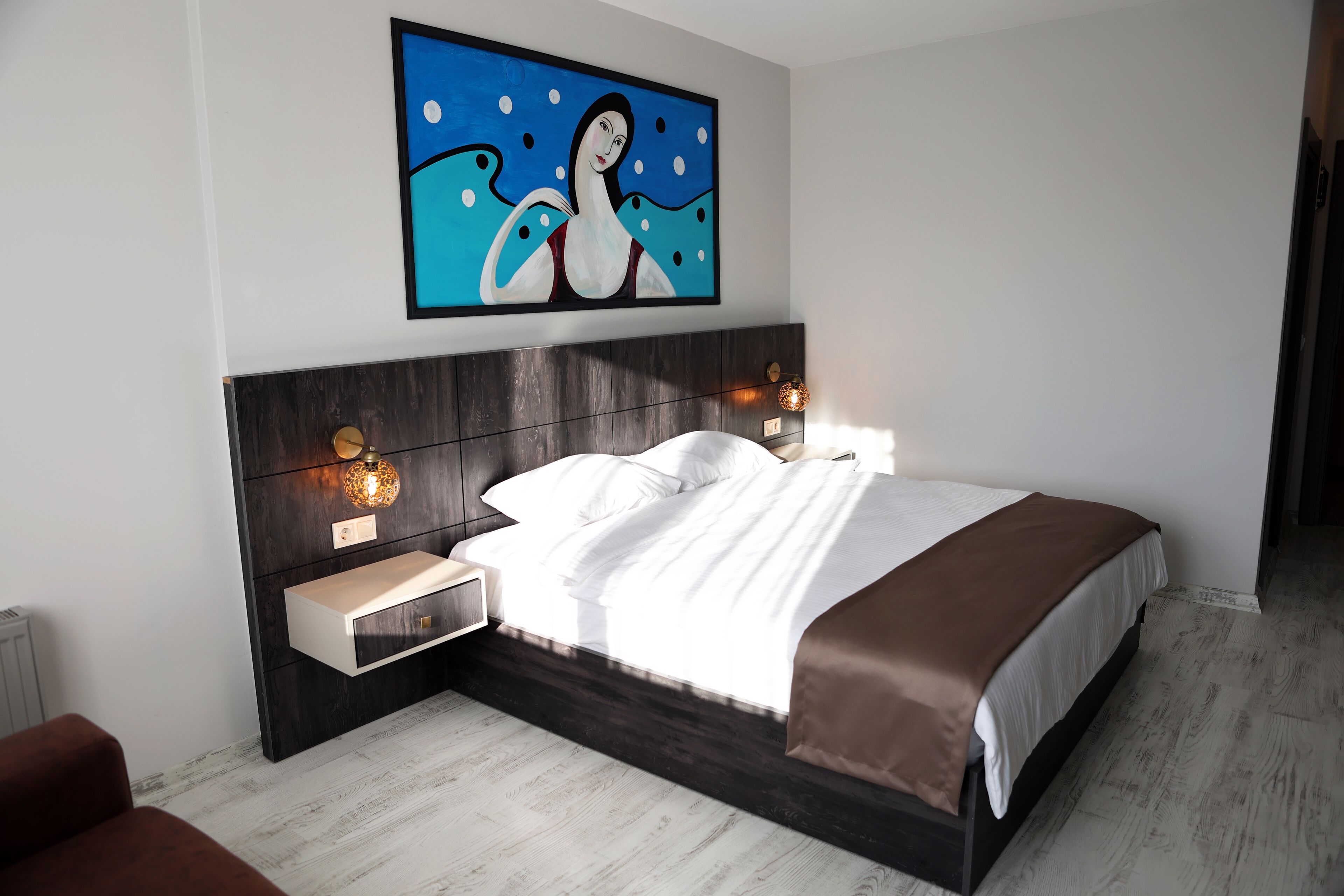 A modern bedroom with a large bed, a nightstand, a wall-mounted lamp, and a painting above the bed.
