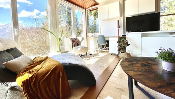 Ein modernes Wohnzimmer mit großem Fenster, einer Couch, einem Schreibtisch und einem Fernseher.