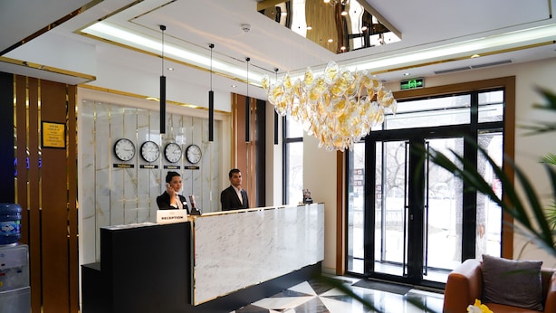 Un hall d’hôtel avec un comptoir de réception en marbre, deux réceptionnistes, un grand lustre et un sol orné de motifs géométriques.