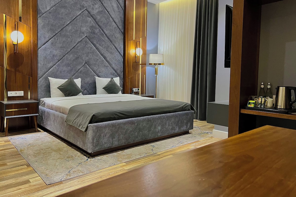 A modern hotel room with a large bed, a wooden nightstand, a desk, and a wardrobe.