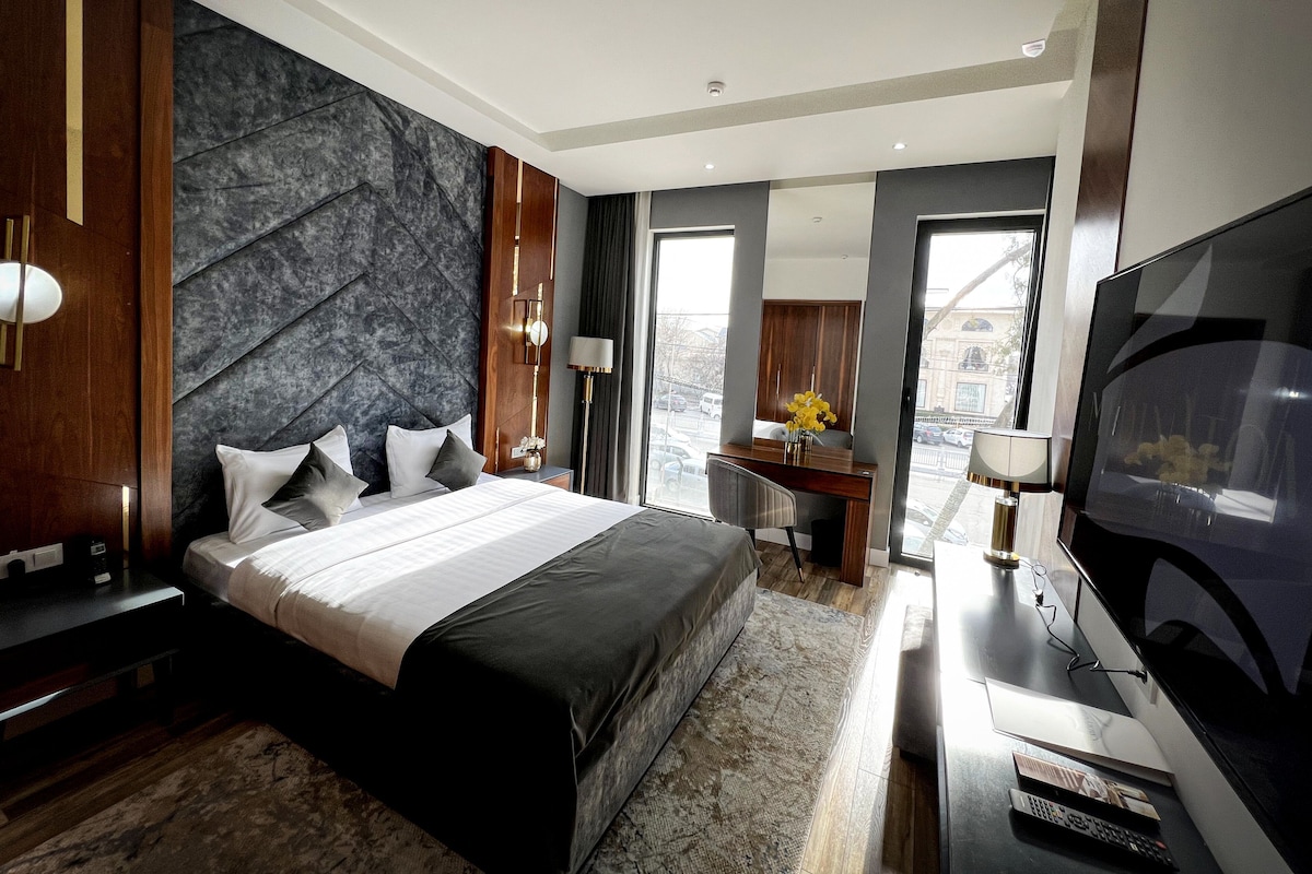 A modern hotel room with a large bed, a desk, a flat-screen TV, and a city view.