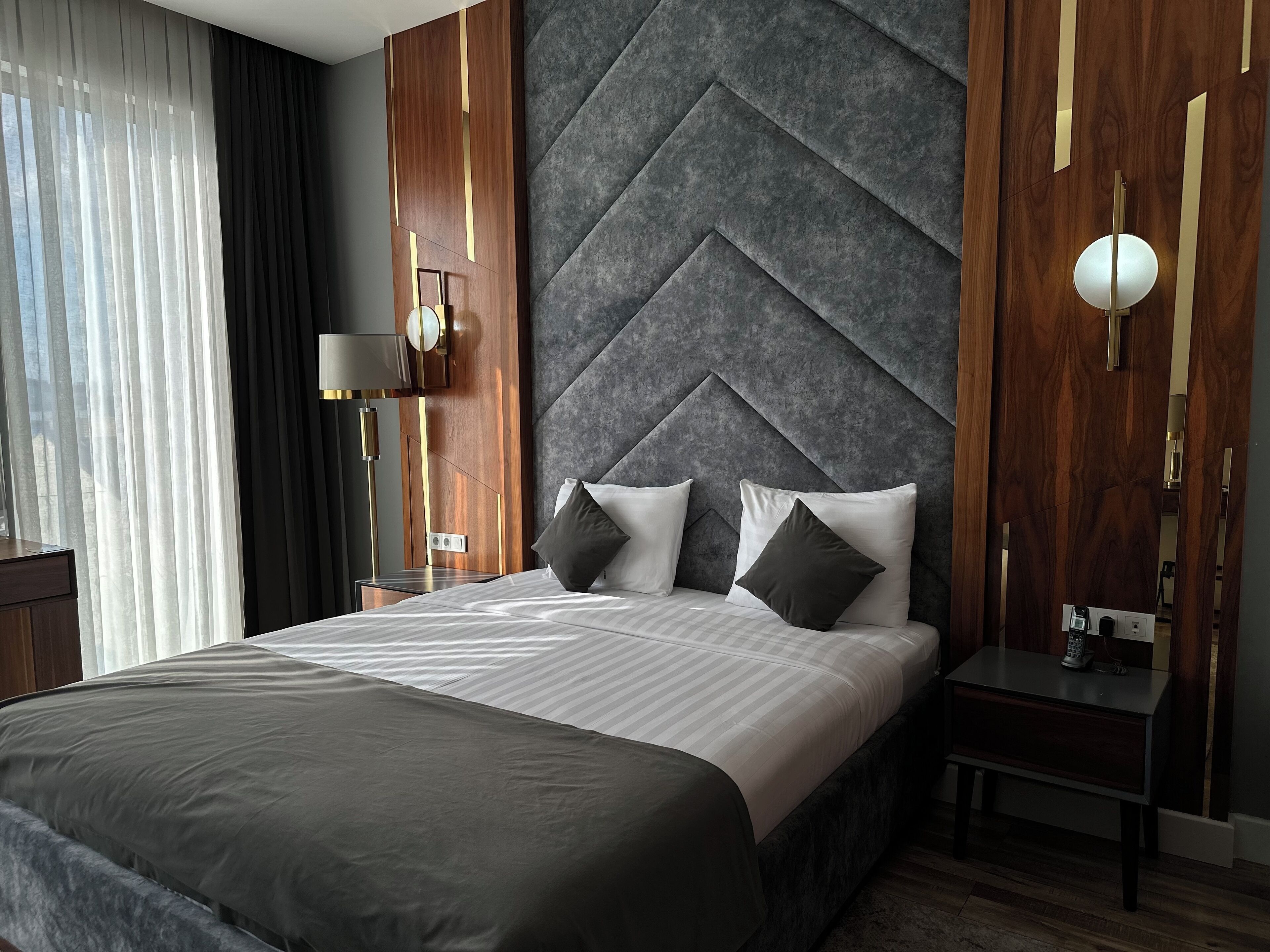 A modern hotel room with a large bed, a nightstand, a lamp, and a view of the outdoors through sheer curtains.