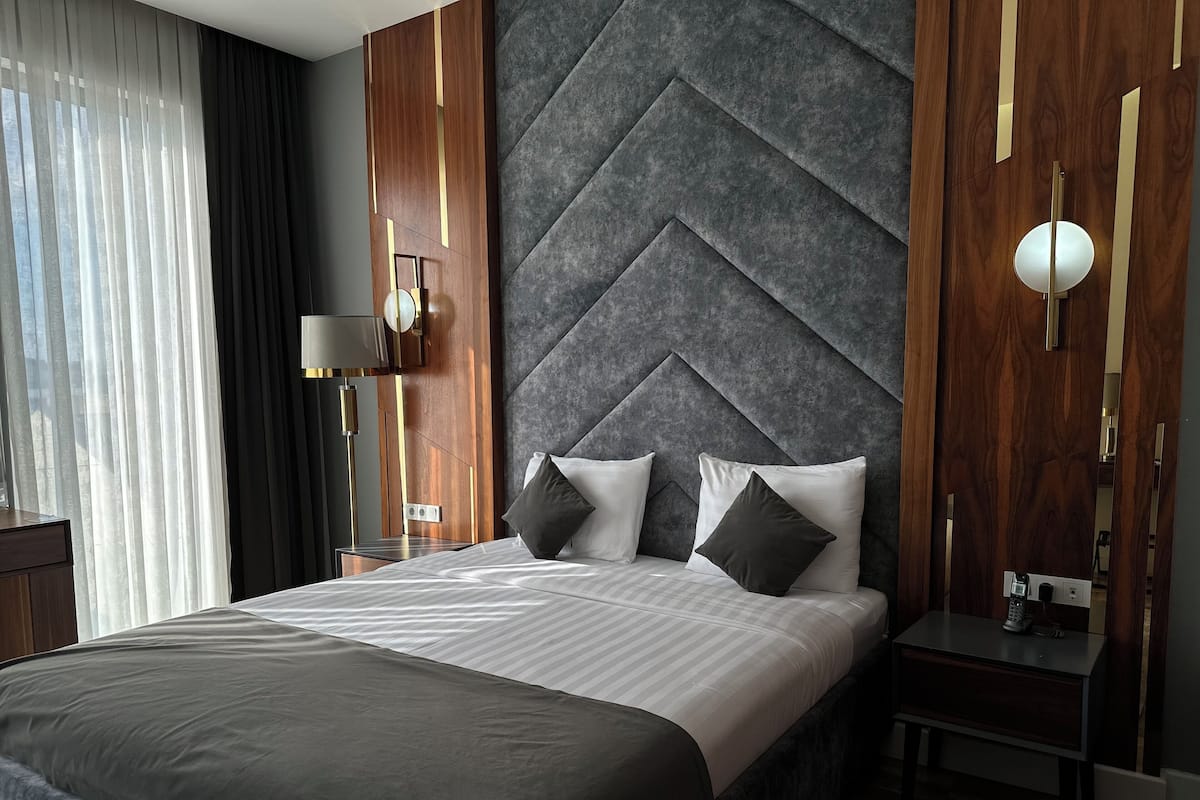 A modern hotel room with a large bed, a nightstand, a lamp, and a view of the outdoors through sheer curtains.