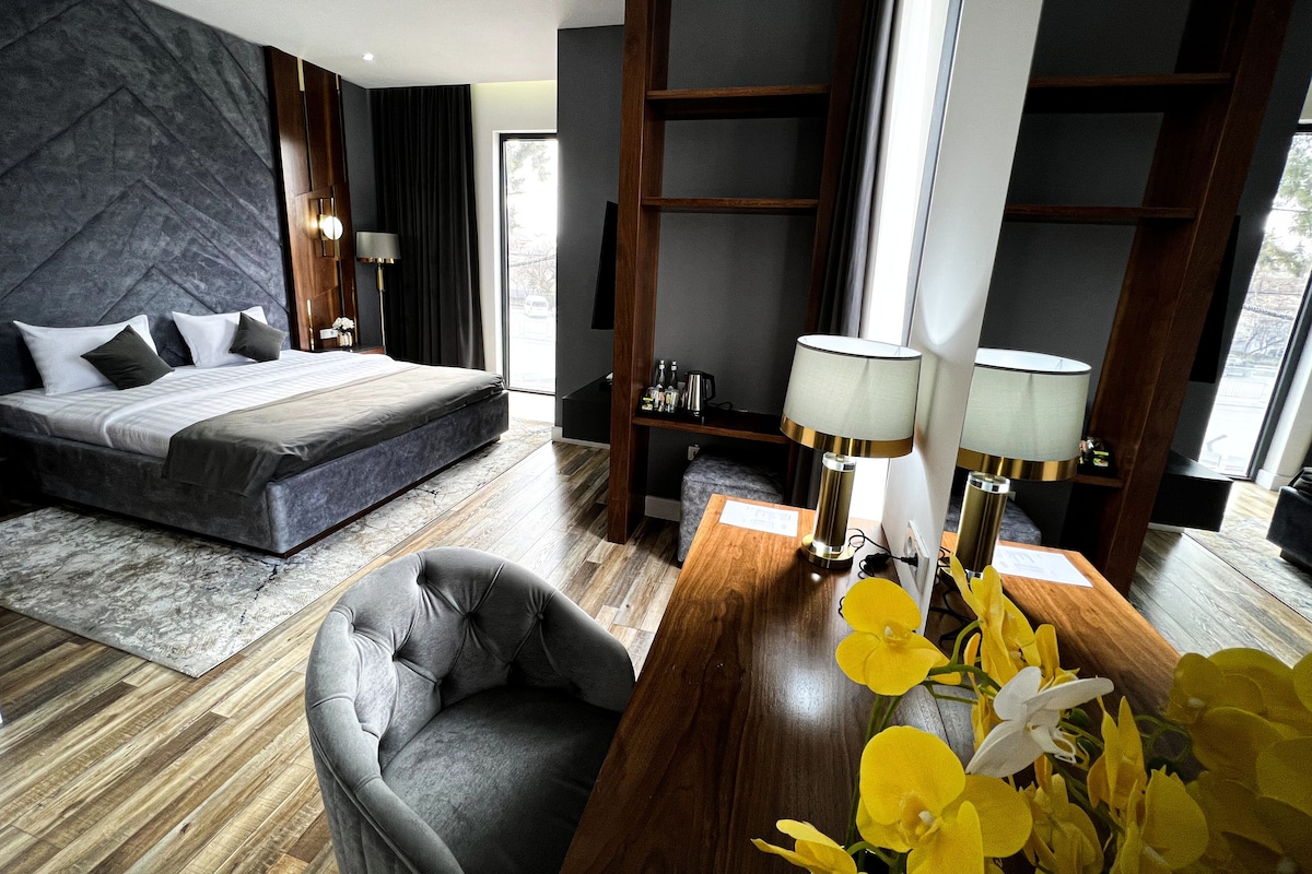 A modern hotel room with a large bed, a seating area, a desk, and a view of the outdoors.