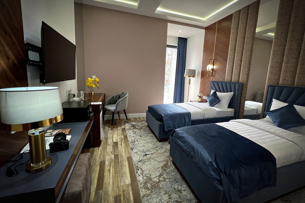 A hotel room with two beds, a desk, a chair, a TV, and a lamp.