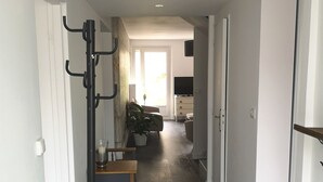 A hallway with a dark wooden floor, white walls, and a ceiling with recessed lighting. There is a coat rack on the left, a small table with a plant, and a television on a stand in the background.
