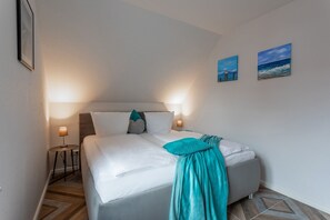 Un lit bien fait, avec du linge de lit blanc et un plaid turquoise, deux lampes de chevet et deux œuvres d’art encadrées accrochées au mur.