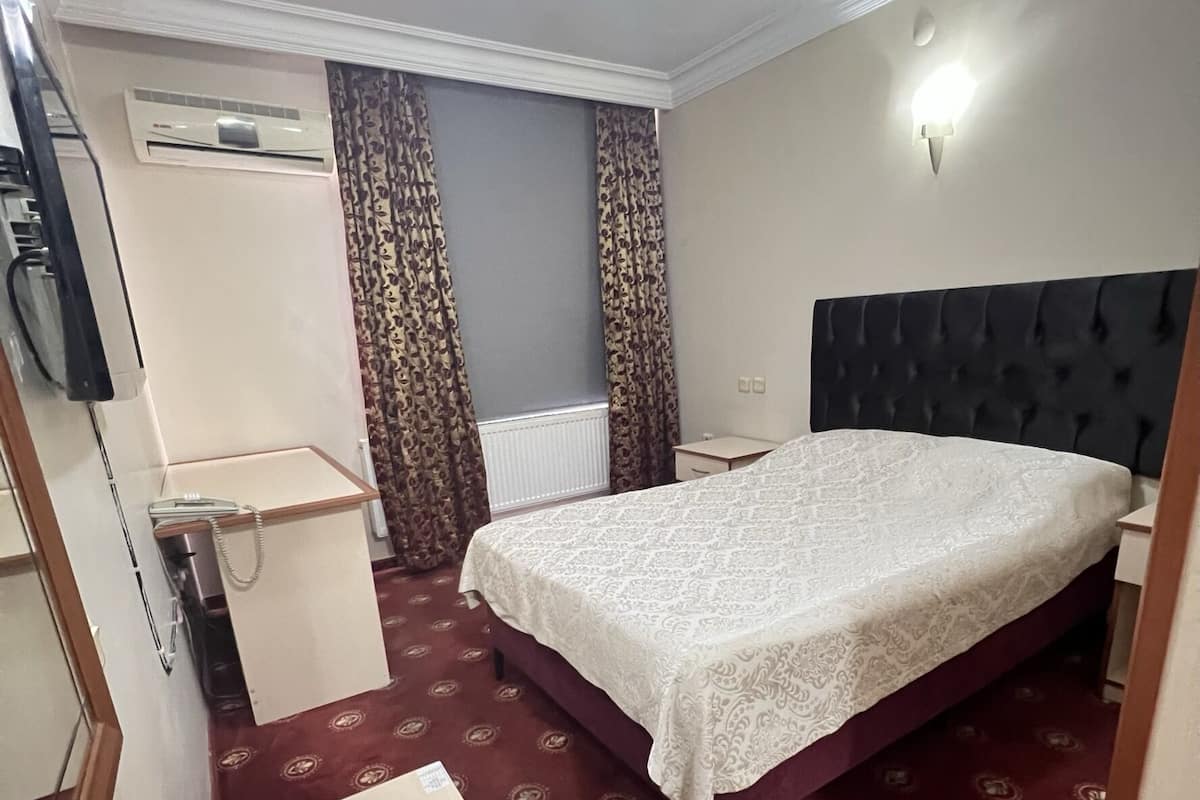A hotel room with a bed, a desk, a chair, a radiator, and curtains.