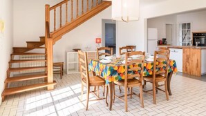 Een ruime eethoek met een houten trap, een tafel met kleurrijk tafelkleed, en een keuken op de achtergrond.