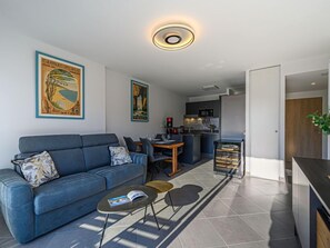 A modern living room with a sofa, coffee table, and dining area. There is a kitchen in the background with a microwave and a coffee machine.