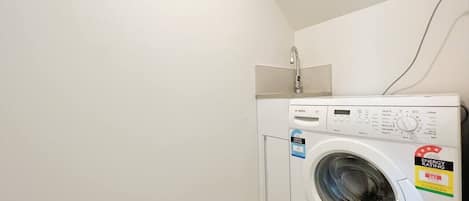 Une petite machine à laver blanche, dans une petite buanderie aux murs blancs et au plafond en pente.