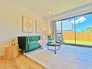 Een moderne woonkamer met een groene bank, een ronde salontafel en een groot raam met uitzicht op een houten schutting.