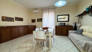 A compact living space with a dining table, chairs, a sofa, a kitchen area with cabinets, a microwave, and a hanging light fixture. - App. Dea Minerva, 3 bedrooms, near the beach (Torre San Giovanni)
