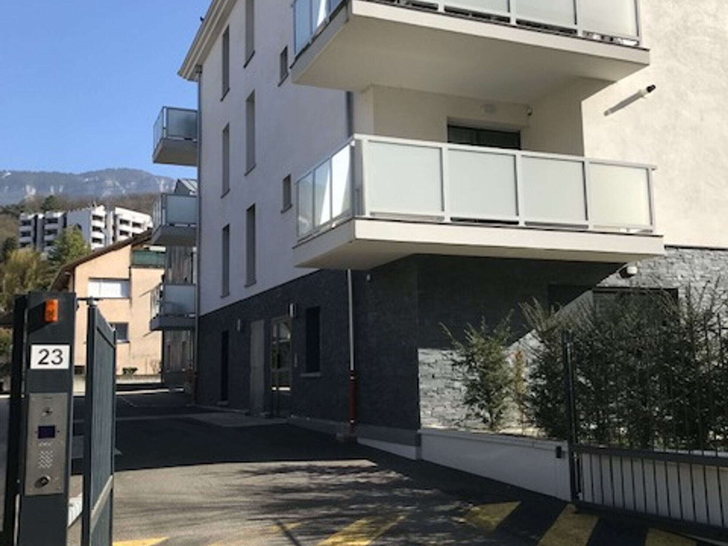 Ein modernes Apartmenthaus mit Balkon, einem mit '23' beschrifteten Tor und freiem Blick auf die Straße sowie Berge im Hintergrund.